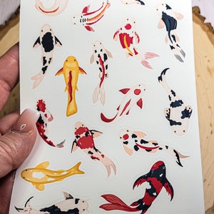 May include: A sheet of stickers featuring 16 colorful koi fish in various poses. The stickers are on a white background with the text "Koi Fish" at the top and "SakuraDesignGallery" at the bottom.