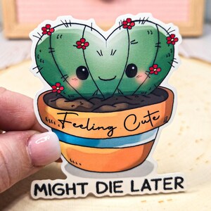 May include: A green heart-shaped cactus in a brown pot with the text "Feeling Cute" and "Might Die Later" on the sticker. The cactus has a cute cartoon face with big eyes and a smile.