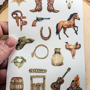 May include: A sheet of stickers with a Western theme. The stickers feature images of a sheriff's badge, cowboy boots, a skull, a horse, a gun, a horseshoe, a cowboy hat, a lasso, a saddle, a bandana, a saloon sign, a swinging saloon door, a guitar, and a campfire.