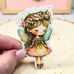May include: A sticker featuring a watercolor illustration of a fairy with green hair, a floral crown, and butterfly wings. The fairy wears a pink and yellow dress and has rosy cheeks. The sticker is held by a hand.