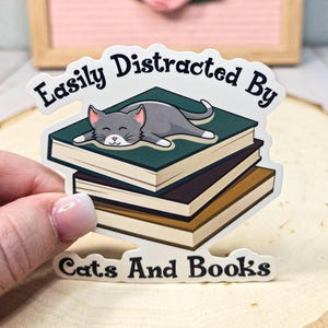 May include: A white sticker with a gray cat sleeping on a stack of books. The sticker says "Easily Distracted By Cats And Books".