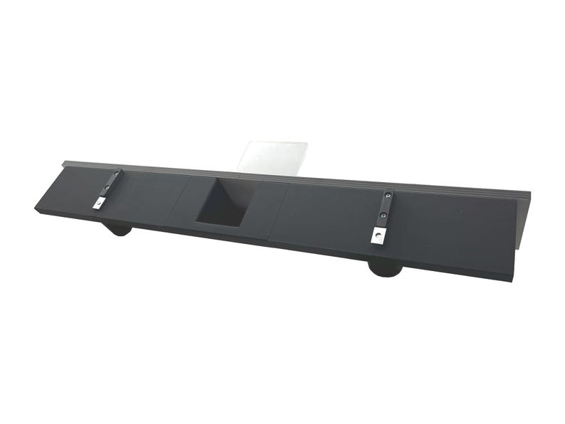 May include: A black, rectangular shelf with a recessed center section and rounded supports. The shelf has two metal brackets attached to the top surface. The shelf is designed to be mounted on a wall.
