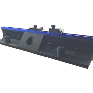 May include: A black and blue metal tool with a rectangular base. The tool has a blue top edge and two black knobs. A circular hole is cut into the center of the tool. The tool is designed for precision work.