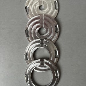 May include: Four silver metal rings with a circular design and a hole in the center. The rings are stacked on top of each other.