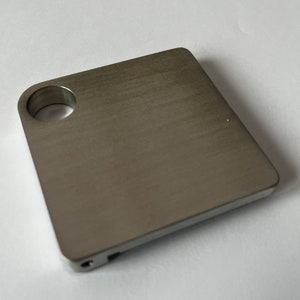 Pode incluir: Um chaveiro quadrado de metal prateado com um furo redondo na parte superior e um pequeno furo na lateral.