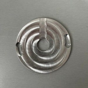 May include: A silver metal circular disc with a concentric circle design and a hole in the center. The disc has a textured surface and two small holes on the side.