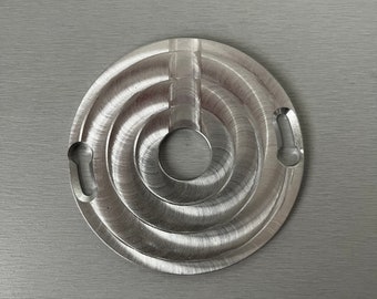 May include: A silver metal circular disc with a concentric circle design and a hole in the center. The disc has a textured surface and two small holes on the side.