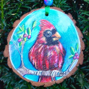 May include: A hand-painted wooden ornament featuring a red cardinal perched on a branch with green leaves and red berries. The background is a bright blue sky. The ornament is attached to a green ribbon with a blue and pink bead.