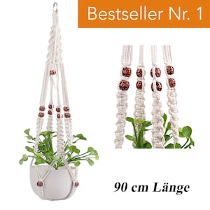 Könnte beinhalten: Cremefarbener Makramee-Pflanzenaufhänger mit einem weißen Keramiktopf, der eine grüne Pflanze enthält. Der Aufhänger hat braune Perlen und einen Metallring. Der Text "Bestseller Nr. 1" ist sichtbar. Die Länge beträgt ca. 90 cm.