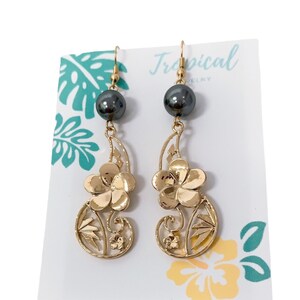 May include: Gold earrings with a black pearl and a floral design. The earrings are on a white card with the word "Tropical" printed on it.