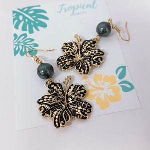 May include: Gold hibiscus flower earrings with black and gold detailing and a dark green pearl dangle. The earrings are on a white background with a card that says "Tropical Jewelry".