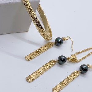 May include: Gold jewelry set with a bangle bracelet, a pendant necklace, and earrings. Each piece features an intricate floral design and is accented with black pearls.