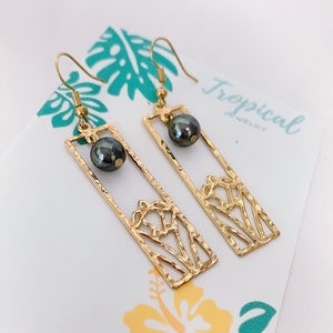 May include: Gold earrings with a black pearl and a cutout design of a tropical flower. The earrings are on a white card with the text "Tropical Jewelry".
