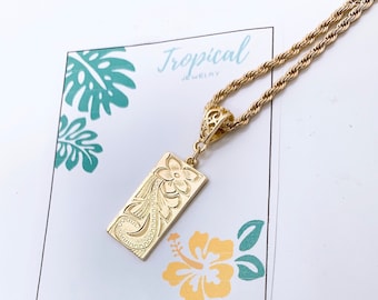 Op de afbeelding: Gouden ketting met een rechthoekige hanger met een bloemmotief. De hanger is bevestigd aan een touwketting. De ketting wordt gepresenteerd op een witte kaart met een groen tropisch bladontwerp en de tekst "Tropical Jewelry".