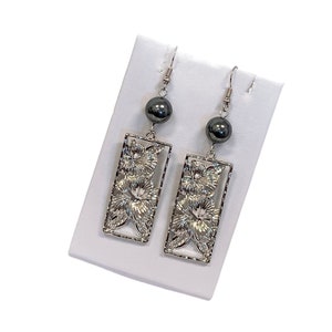 May include: Silver earrings with a rectangular floral design and a black bead dangle.