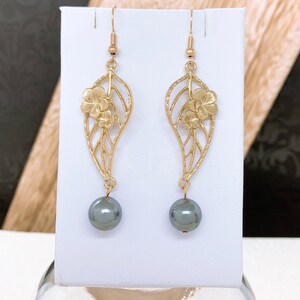 Peut inclure: Boucles d'oreilles pendantes dor&eacute;es avec un motif de feuille et un petit d&eacute;tail de fleur. Les boucles d'oreilles ont une perle noire.
