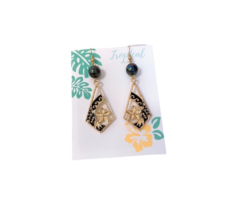 May include: Gold earrings with black and gold floral designs and black pearl accents. The earrings are displayed on a white card with the word "Tropical" printed on it.