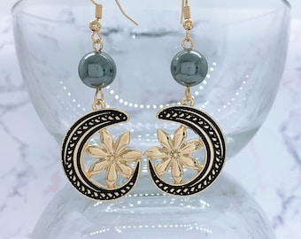 May include: A pair of gold earrings with a crescent moon design and a black and gold flower accent. The earrings feature a dark gray bead.