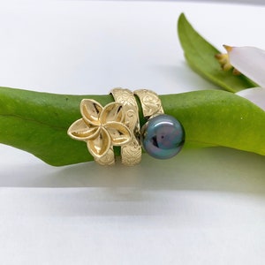 May include: A gold ring with a flower design and a black pearl. The ring is on a green leaf.