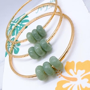 May include: Three gold-colored bangles with a hammered texture, each adorned with four light green jade beads. The bangles rest on a white card with a floral design and the text "Tropical Jewel".