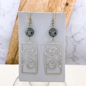 May include: Silver earrings with a wave design and a black pearl dangle. The earrings are displayed on a white background.