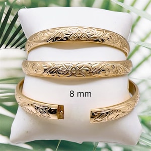 May include: Gold-colored bangles with an engraved floral pattern, displayed on a white cushion. The bangles are 8 mm wide. The open bangle shows the clasp. The background has green foliage.