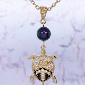 Puede incluir: Collar de cadena dorada con una cuenta negra iridiscente y un colgante de tortuga dorada. La tortuga tiene un dise&ntilde;o detallado con una flor en el centro.