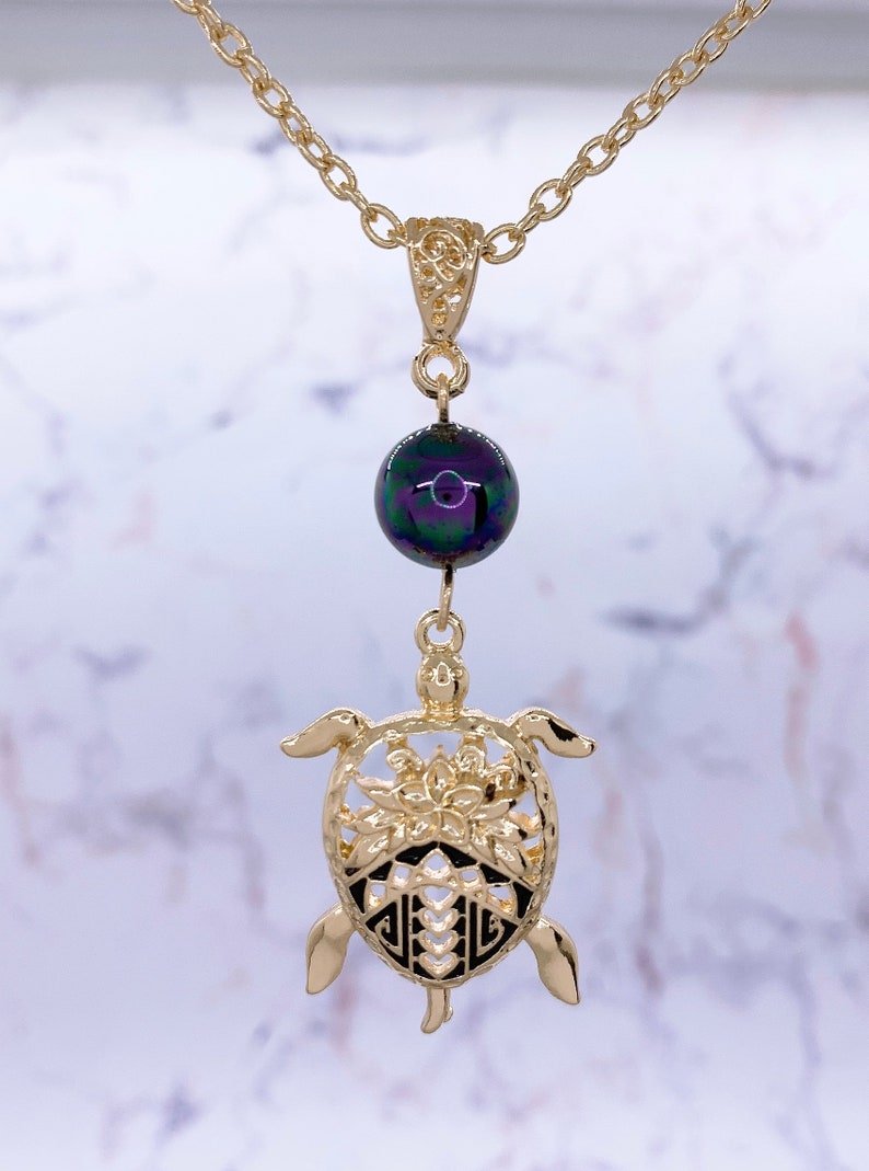 Puede incluir: Collar de cadena dorada con una cuenta negra iridiscente y un colgante de tortuga dorada. La tortuga tiene un dise&ntilde;o detallado con una flor en el centro.