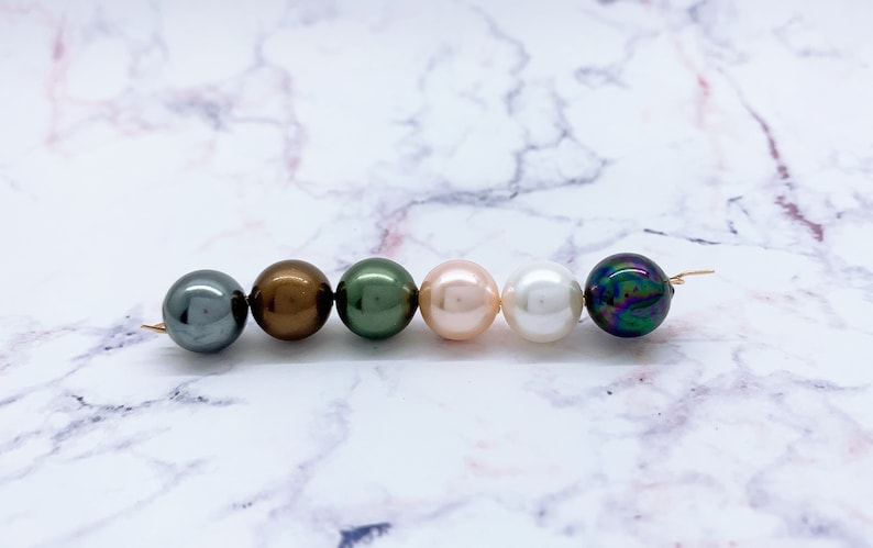 May include: Six round beads in a row on a white marble surface. The beads are various colors, including black, brown, green, pink, white, and iridescent.