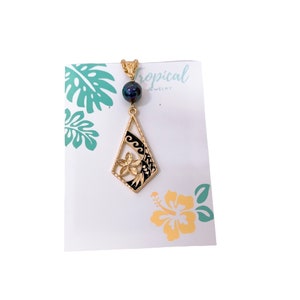 May include: Gold pendant necklace with a black and gold flower design and a black pearl. The pendant is shaped like a diamond and has a wave pattern on the side. The necklace is on a white card with the text "Tropical Jewelry".