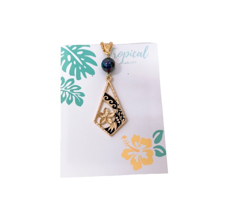 May include: Gold pendant necklace with a black and gold flower design and a black pearl. The pendant is shaped like a diamond and has a wave pattern on the side. The necklace is on a white card with the text "Tropical Jewelry".