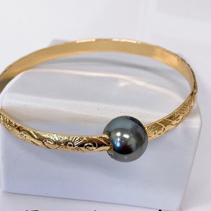 May include: A gold bangle bracelet with an intricate floral design and a dark gray pearl accent.
