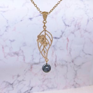 Peut inclure: Un collier en or avec un pendentif d&eacute;licat en forme de feuille orn&eacute; d'un petit motif floral. Une perle noire pend au bas du pendentif.
