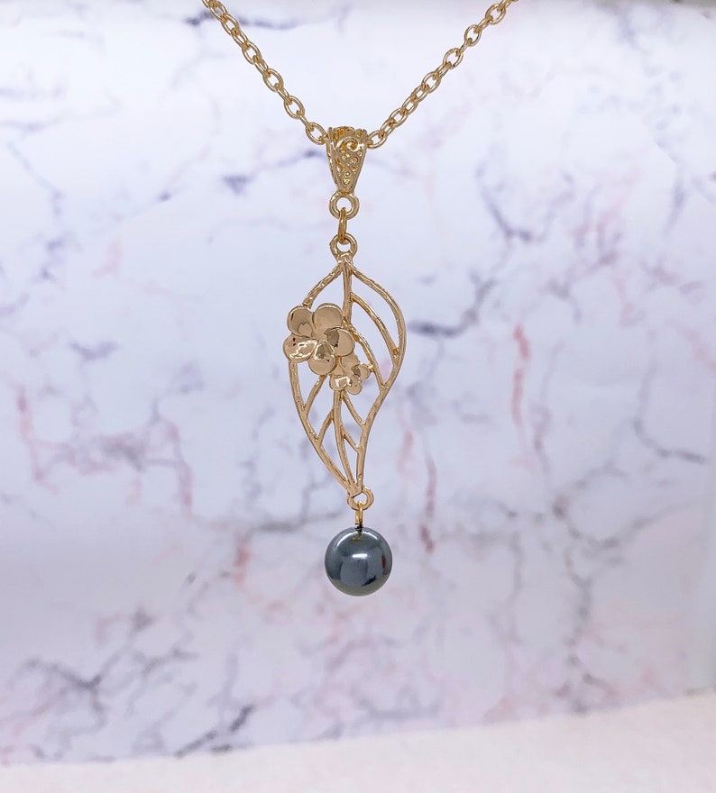 Peut inclure: Un collier en or avec un pendentif d&eacute;licat en forme de feuille orn&eacute; d'un petit motif floral. Une perle noire pend au bas du pendentif.