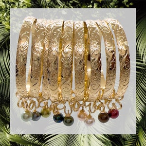 May include: A set of eight gold-tone bangle bracelets with intricate floral designs and dangling charms. The charms are various colors, including green, brown, black, and pink.