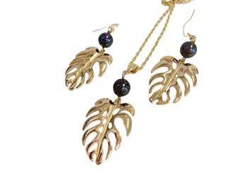 May include: Gold necklace and earrings set with black iridescent beads and gold leaf charms. The leaves are a tropical plant design.