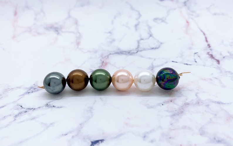 May include: Six round beads in a row on a white marble surface. The beads are various colors, including black, brown, green, pink, white, and iridescent.