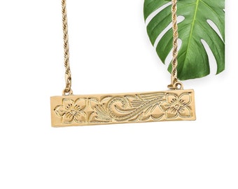 Op de afbeelding: Een gouden ketting met een touwketting en een rechthoekige hanger met een bloemenmotief.