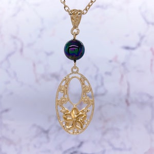 K&ouml;nnte beinhalten: Eine goldene Halskette mit einem zarten ovalen Anh&auml;nger mit einem floralen Design und einer einzigen schillernden schwarzen Perle.