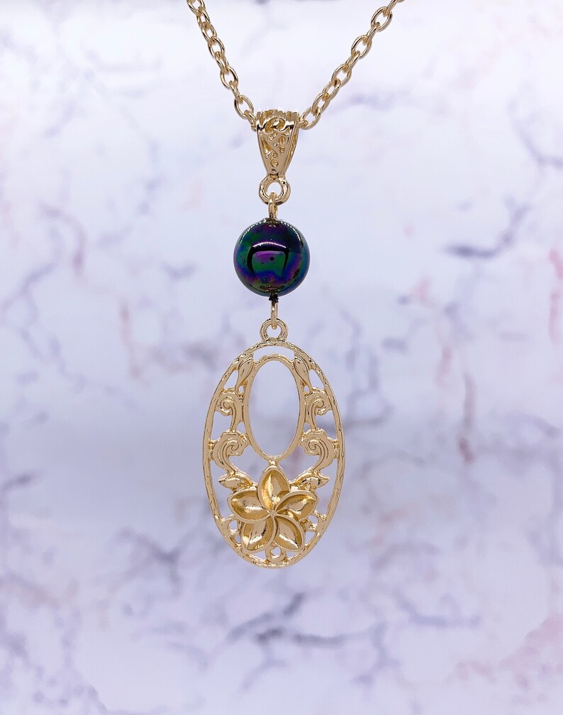 K&ouml;nnte beinhalten: Eine goldene Halskette mit einem zarten ovalen Anh&auml;nger mit einem floralen Design und einer einzigen schillernden schwarzen Perle.