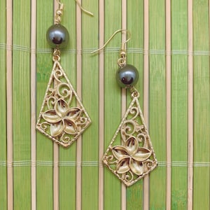 May include: A pair of gold earrings with black pearl accents and a floral design. The earrings are hanging from a bamboo mat background.