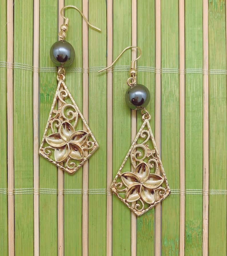 May include: A pair of gold earrings with black pearl accents and a floral design. The earrings are hanging from a bamboo mat background.