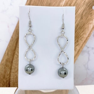 May include: Silver infinity symbol earrings with a dark gray pearl dangling from each earring.
