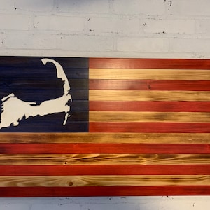 May include: A wooden American flag with a white outline of Cape Cod on the blue field. The flag is made of reclaimed wood and has a rustic, distressed look.