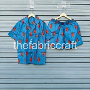 May include: Blue pajama set with a red lobster pattern. The set includes a short-sleeved button-down shirt and matching shorts. The shirt and shorts have white trim. The text "thefabriccraft" is visible on the bottom of the image.