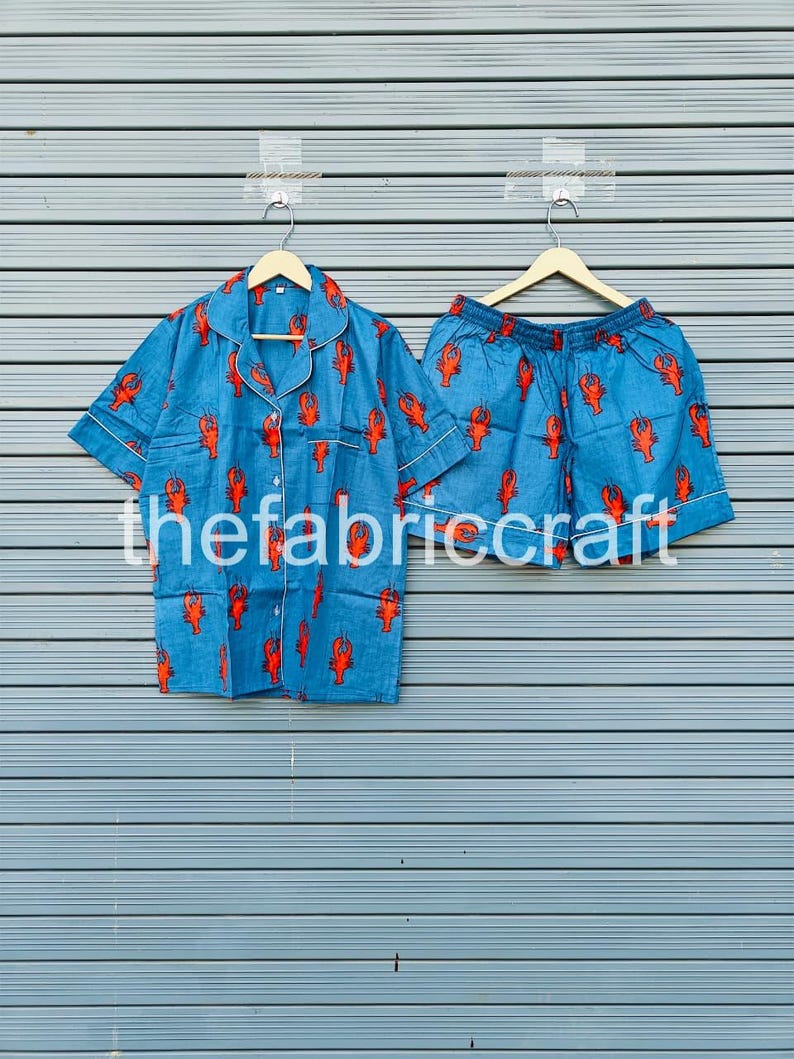 May include: Blue pajama set with a red lobster pattern. The set includes a short-sleeved button-down shirt and matching shorts. The shirt and shorts have white trim. The text "thefabriccraft" is visible on the bottom of the image.