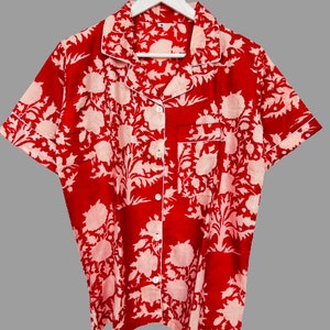 May include: Red short-sleeved button-up shirt with a white floral print. The shirt has a collared neckline and a pocket on the left chest.
