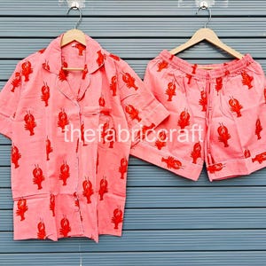 May include: A two-piece pajama set featuring a light pink button-down shirt and matching shorts. The set is adorned with a repeating pattern of red lobsters. The shirt has a chest pocket and white piping.