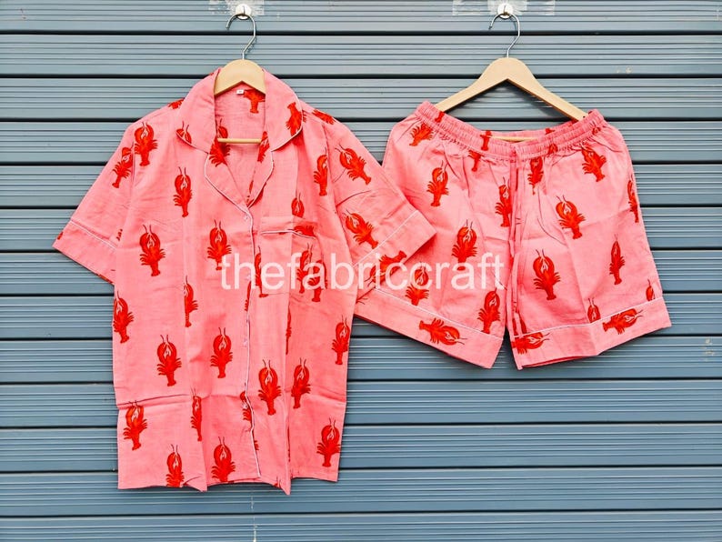 May include: A two-piece pajama set featuring a light pink button-down shirt and matching shorts. The set is adorned with a repeating pattern of red lobsters. The shirt has a chest pocket and white piping.