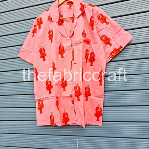 May include: Pink pajama set with a red lobster print. The short-sleeved top has a notched collar, a chest pocket, and white trim. The fabric is hanging on a wooden hanger.